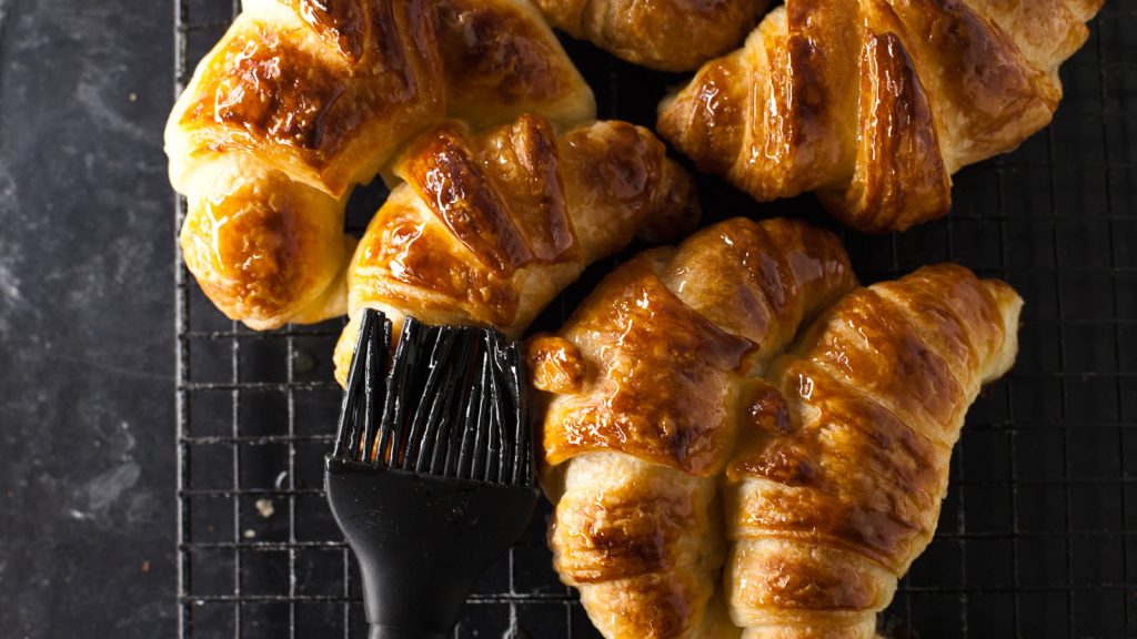 Medialunas caseras de manteca sobre rejilla siendo pintadas con almíbar con pincel de silicona
